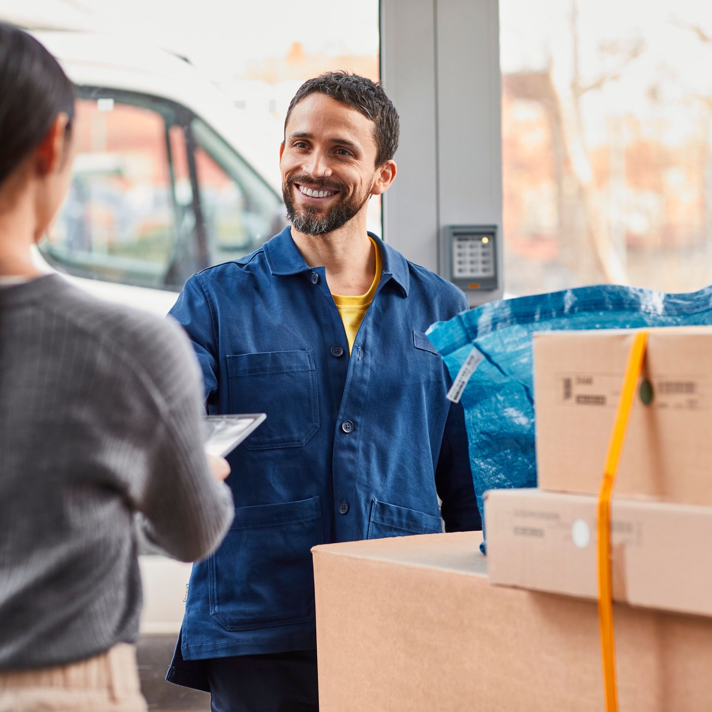 A person in a blue shirt is standing next to flatpack and a blue IKEA bag, talking to another person in a grey sweater.