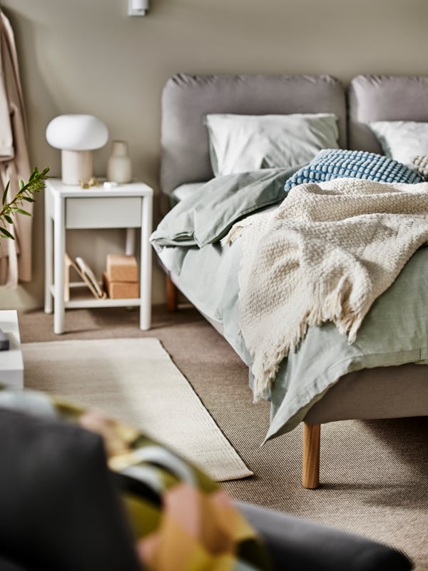 An upholstered bed with green/stripe BERGPALM bed linen and a LAPTÅG throw on it, and a TONSTAD bedside table beside it.