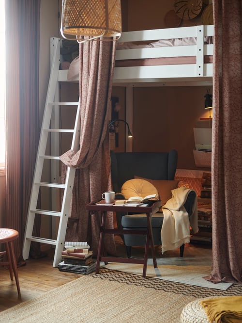 A white loft bed with a cosy reading corner underneath with an armchair, tray table, soft textiles and mood lighting.