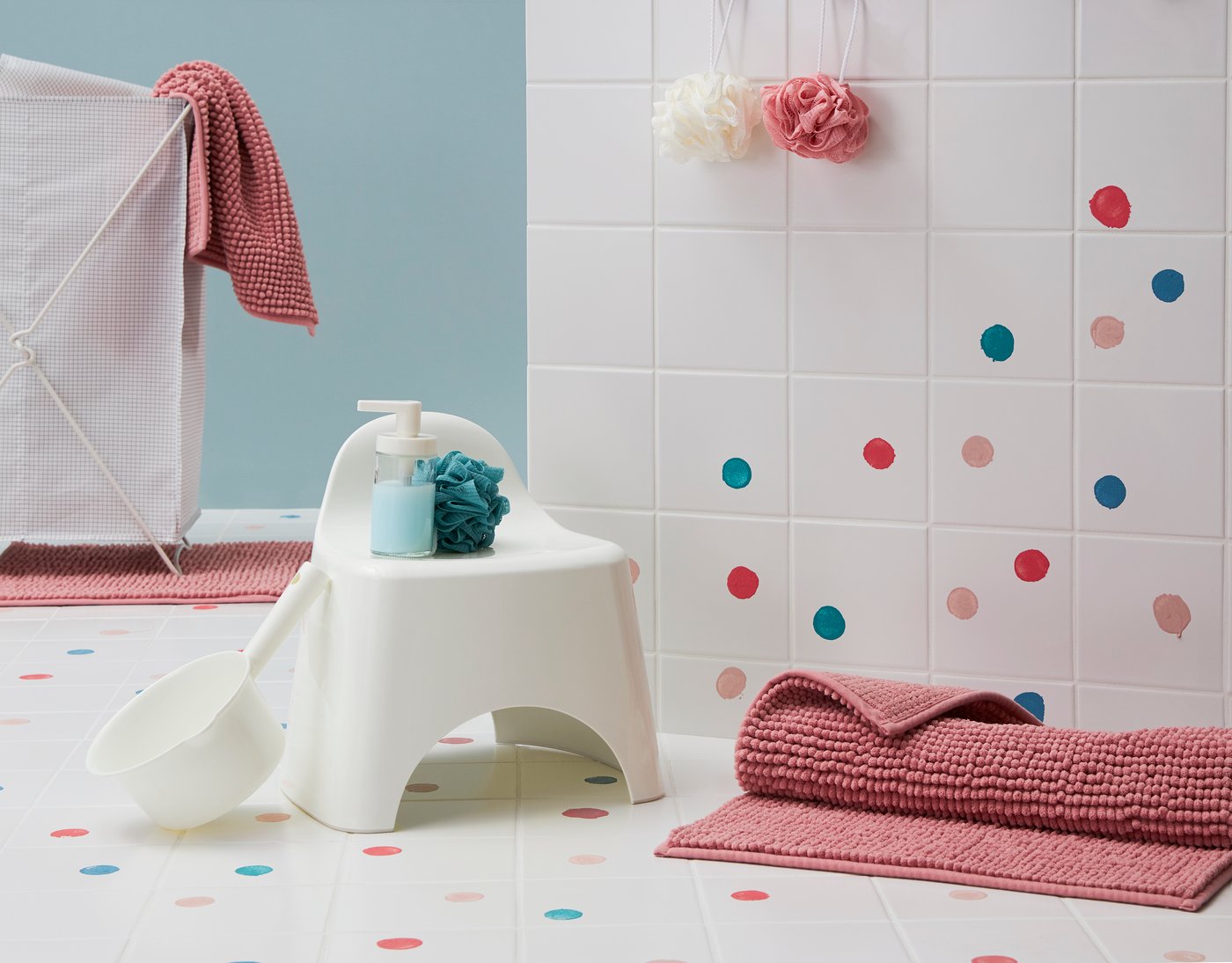 Tapis de bain rose et marche pied en plastique blanc dans une salle de bain blanche avec des pastilles de couleur collées sur le carrelage.