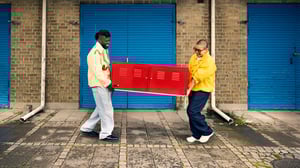 Dwie osoby stoją na chodniku przed budynkiem z niebieskimi drzwiami garażowymi, wspólnie niosąc czerwony metalowy mebel z drzwiczkami. W tle ceglana ściana i rury spustowe, scena wygląda jak przeprowadzka lub transport.