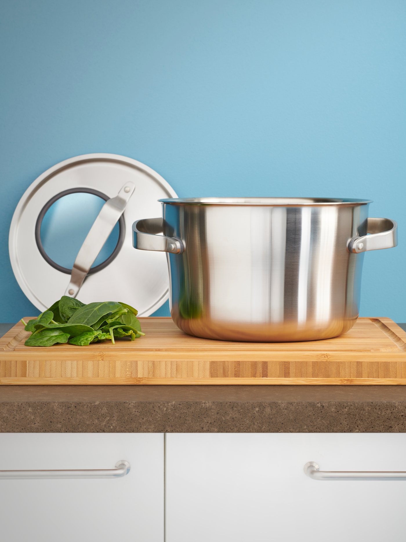 Garnek z serii SENSUELL stoii na  drewnianej desce do krojenia, obok widać pokrywkę opartą o niebieską ścianę.