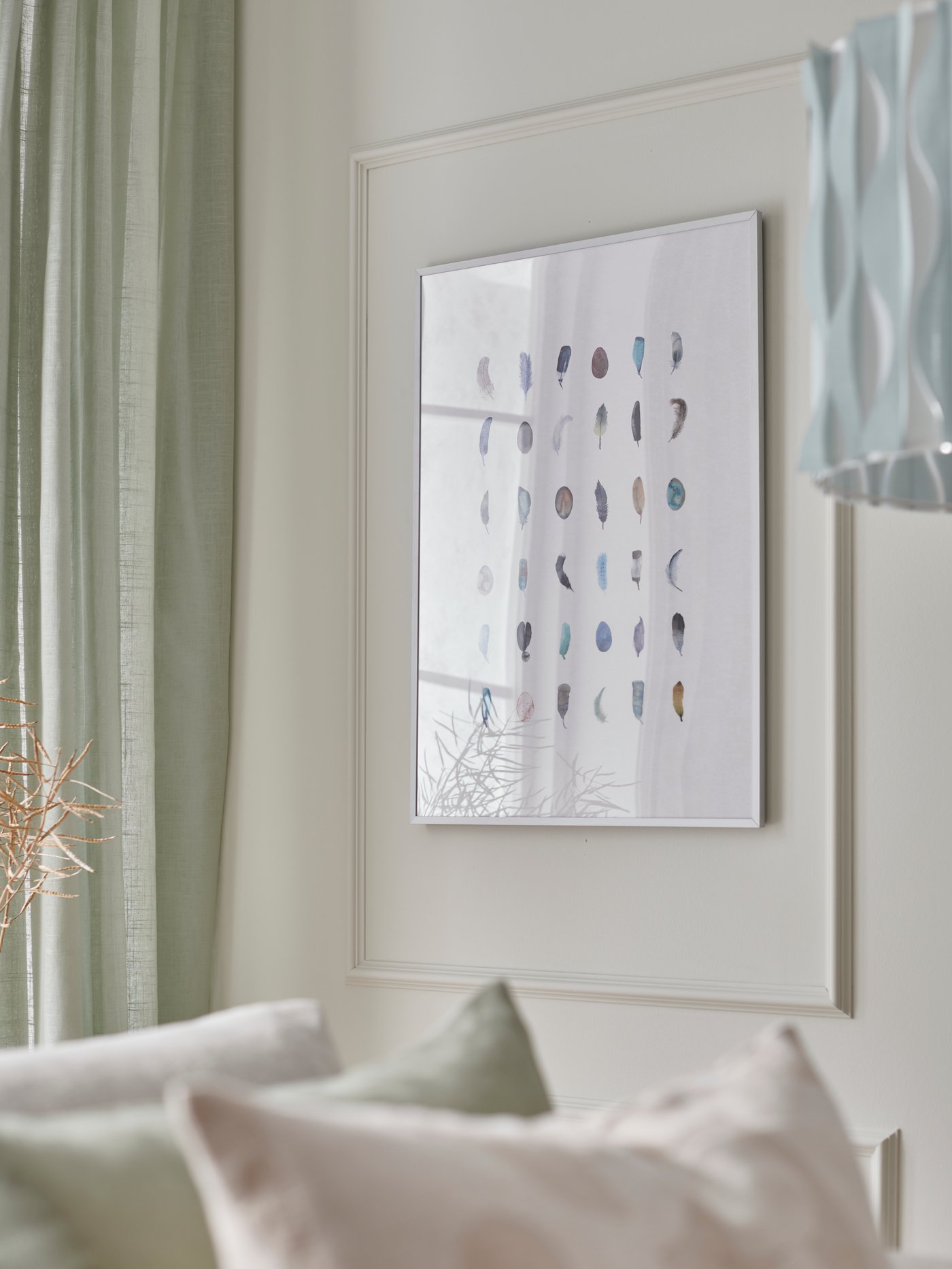 A BILD poster of colourful feathers in a silver LOMVIKEN frame hangs on a cream-coloured wall in a bright living room.