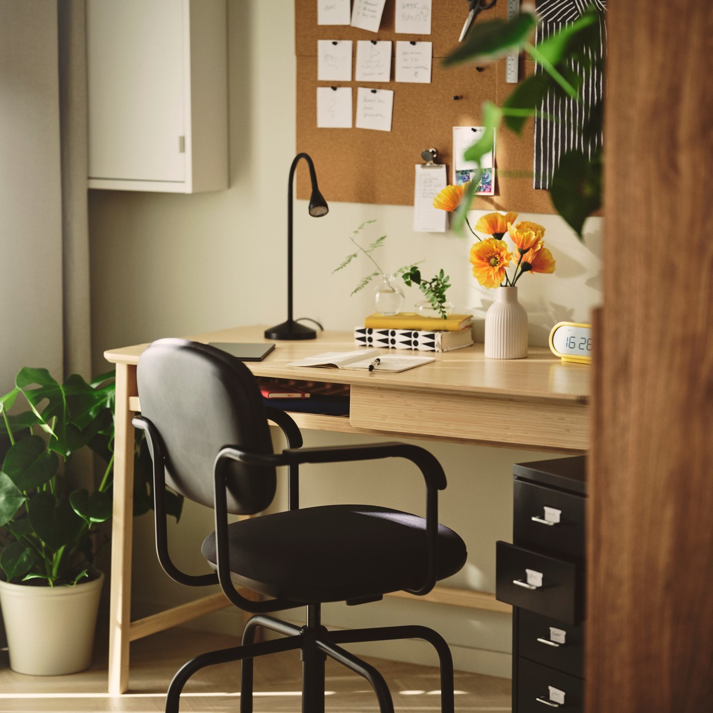 A home office with a dark grey MULLSJÖ swivel chair beside a light wooden desk and a cork board on the wall.