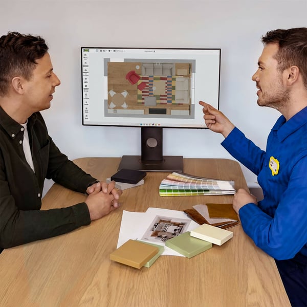 Projektant IKEA siedzący przy biurku w niebieskiej koszulce i omawiający z klientem projekt pokoju wyświetlony na ekranie komputera. Przy biurku siedzi również klient słuchający o rozwiązaniu.