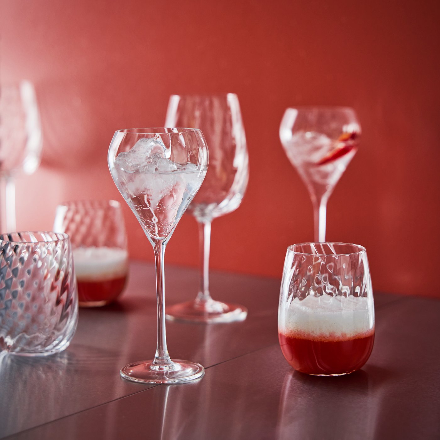 Several FJÄLLUGGLA glasses and champagne glasses filled with drinks stand on a glossy surface against a red background.