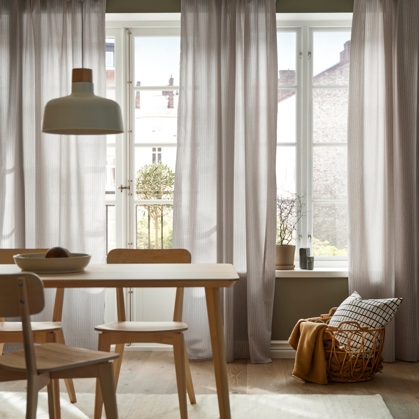 Lys beige gardiner i et rom med et spisebord og tre spisestoler og en kurv som oppbevaring med puter og pledd.