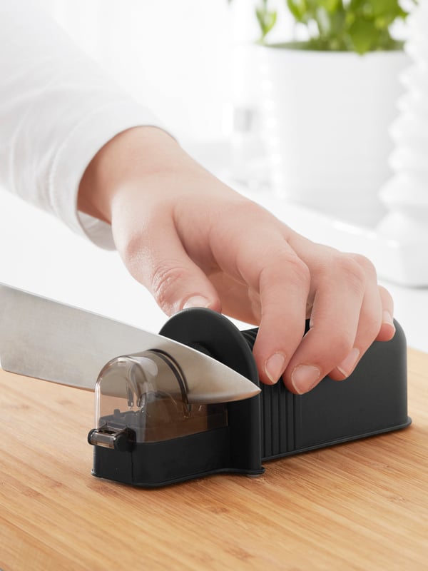 The hand of a person wearing a white long-sleeved top is sharpening a chef’s knife with a black knife sharpener.