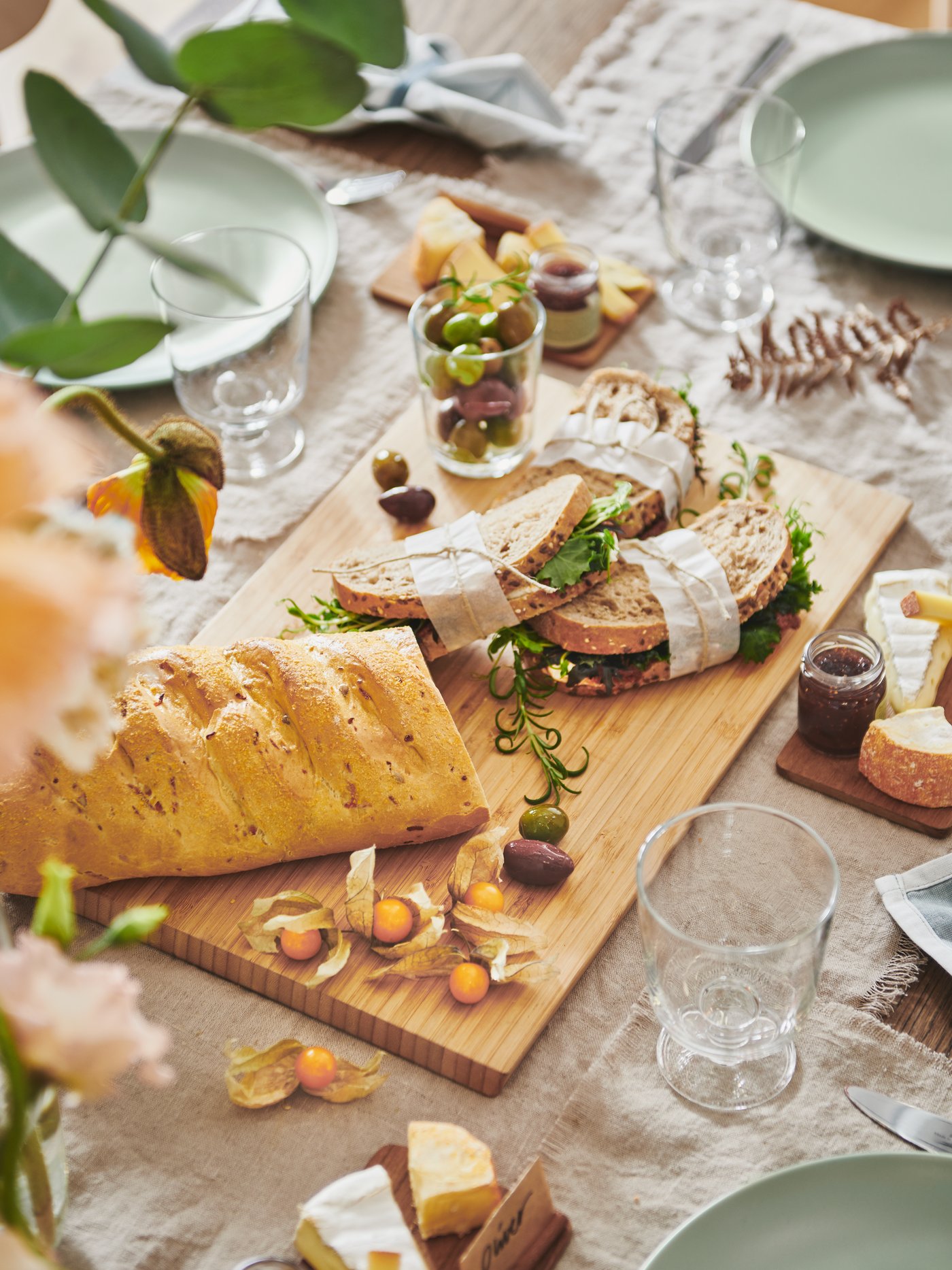 Detalle de una mesa de comedor con una tabla de cortar de madera, platos verdes y copas de vidrio transparente sobre una tela beige.