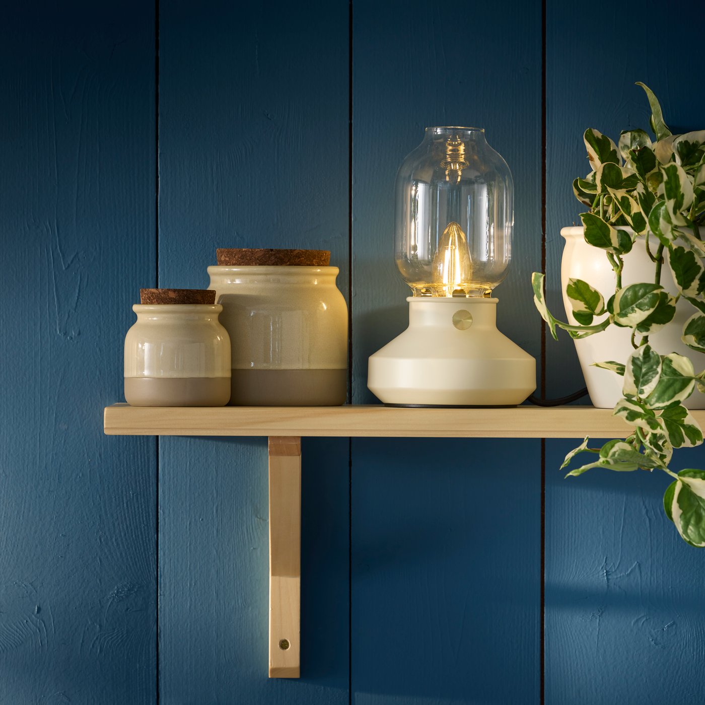 A TÄRNABY table lamp is displayed on a light wooden shelf against a blue wall, accompanied by some jars and a potted plant.