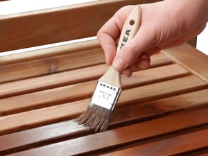 A close-up of a hand holding a paintbrush applying a clear finish to a wooden surface. The bristles of the brush are in contact with the wood, spreading the liquid evenly. The wooden surface has multiple slats, indicating it may be part of furniture or flooring.