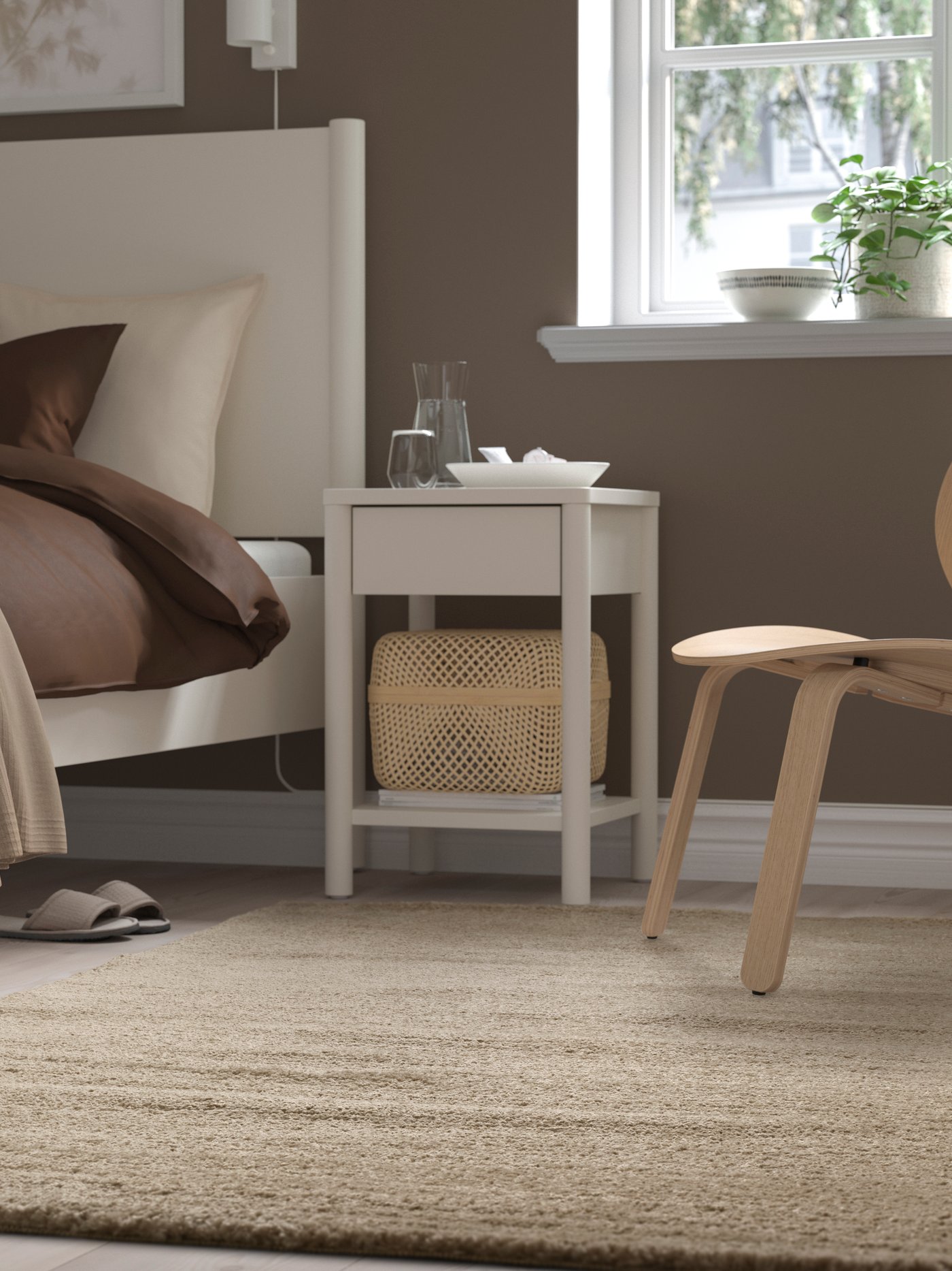 Ein beiger STOENSE Teppich liegt vor einem weißen Einzelbett mit brauner Bettwäsche. Daneben befindet sich ein weißer Ablagetisch und ein FRÖSET Sessel in Eiche.