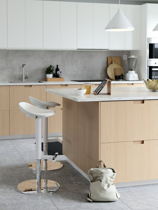 Un cucina in stile scandinavo moderno sui toni naturali del bianco e del legno. Al centro, un'isola con frontali METOD in bambù chiaro e accanto degli sgabelli JANINGE. Sul balcone sono appoggiati un supporto per tablet VIVALLA, un ciotola, un bicchierie e un recipiente con della frutta. Sul pavimento è posato uno zaino beige chiaro. 