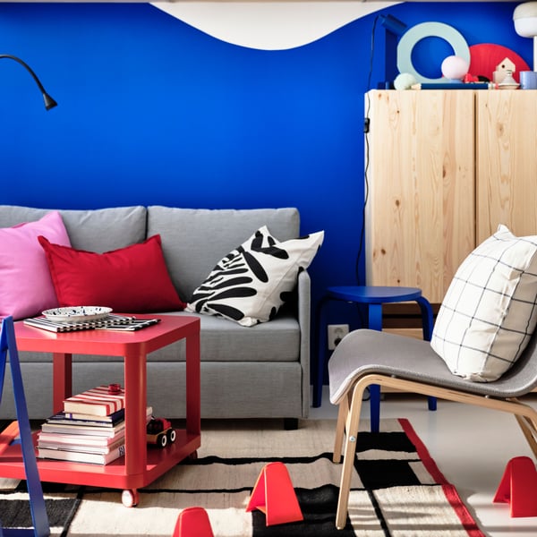 A red TINGBY side table on castors and a NOLMYRA easy chair stand near a FRIDHULT sofa-bed in front of a blue and white wall.