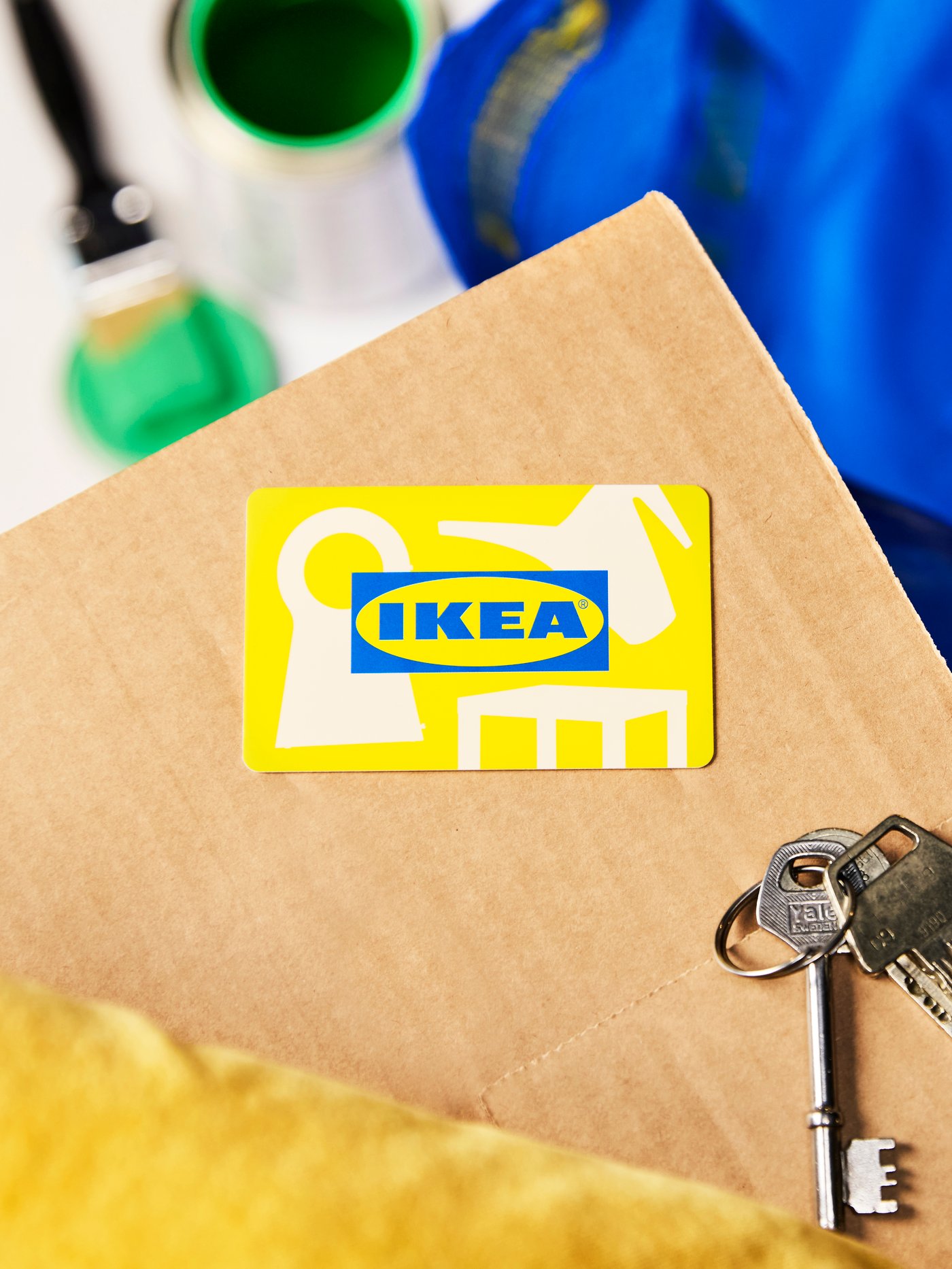 Nahaufnahme einer gelben IKEA Family Karte, die auf einem Umzugskarton neben einem Schlüsselbund liegt – im Hintergrund Farbe, Pinsel und IKEA Tasche