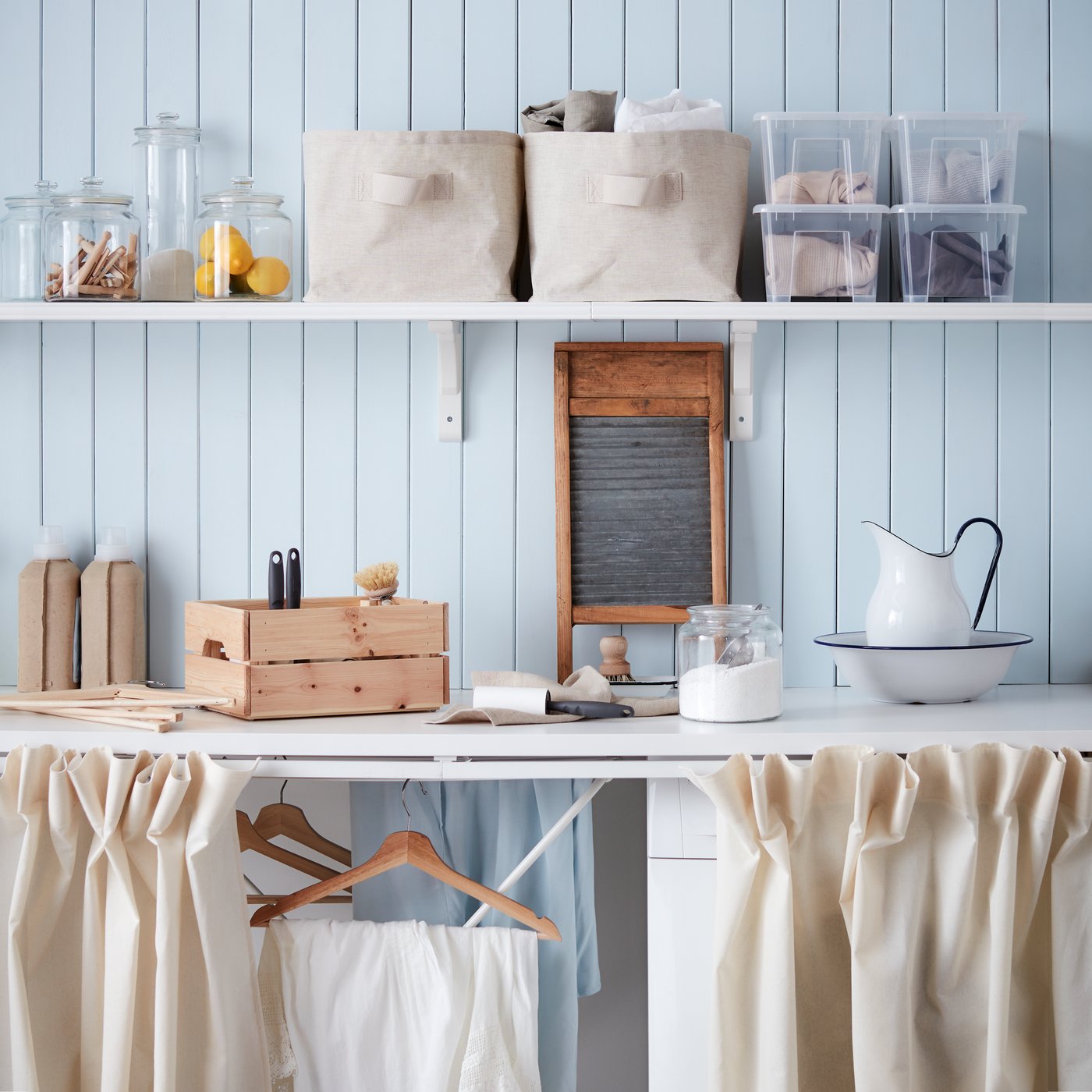 Una tabla de planchar pequeña JÄLL sobre una mesa de cocina blanca con una camisa de jeans por planchar. En una silla de madera frente a la mesa se ve un canasto TORKIS celeste con ropa blanca arrugada. 