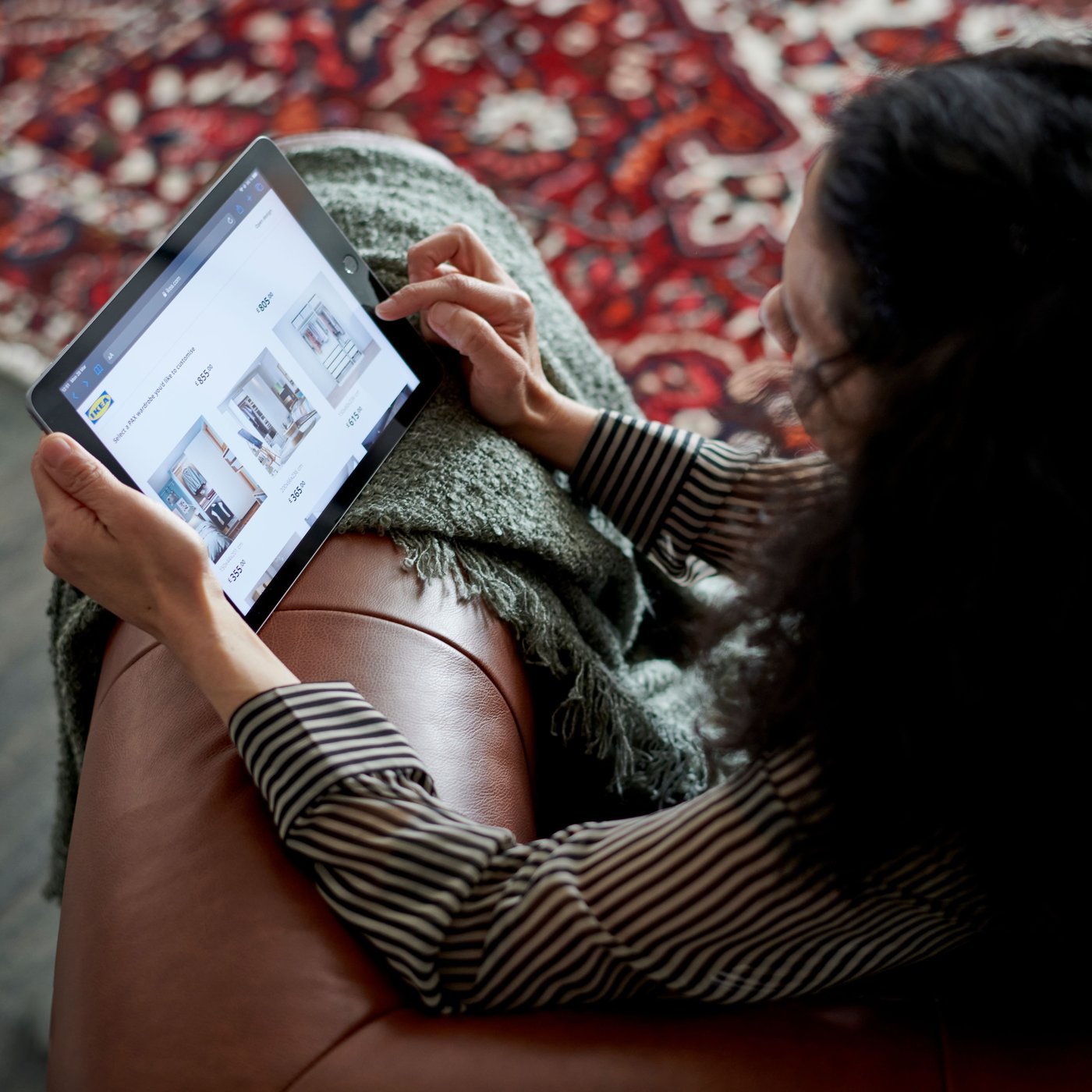 A person sits on a brown sofa with a green throw over the arm. They rest a tablet on the arm and browse the IKEA.com website.