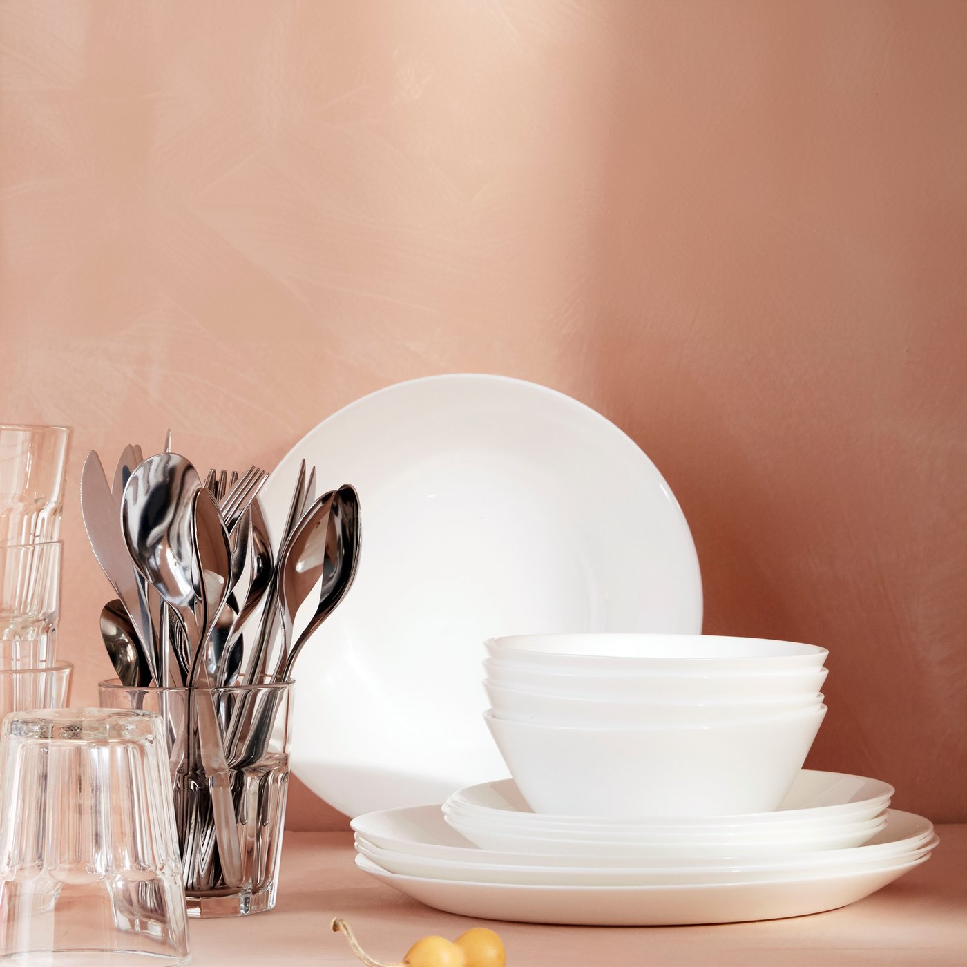 Weißes OFTAST Geschirr-Set mit gestapelten Tellern und Schalen, daneben Gläser und ein Becher mit Besteck vor roséfarbener Wand – schlicht, zeitlos, alltagsbereit.