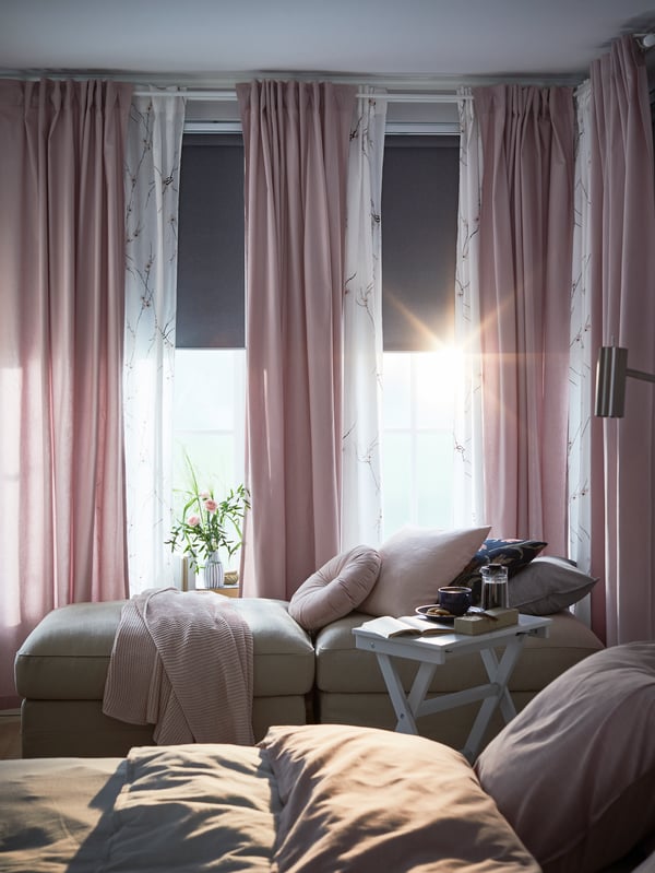 Grey block-out roller blinds half down, pink and white sheer curtains, sofa modules by the window and a white tray table.