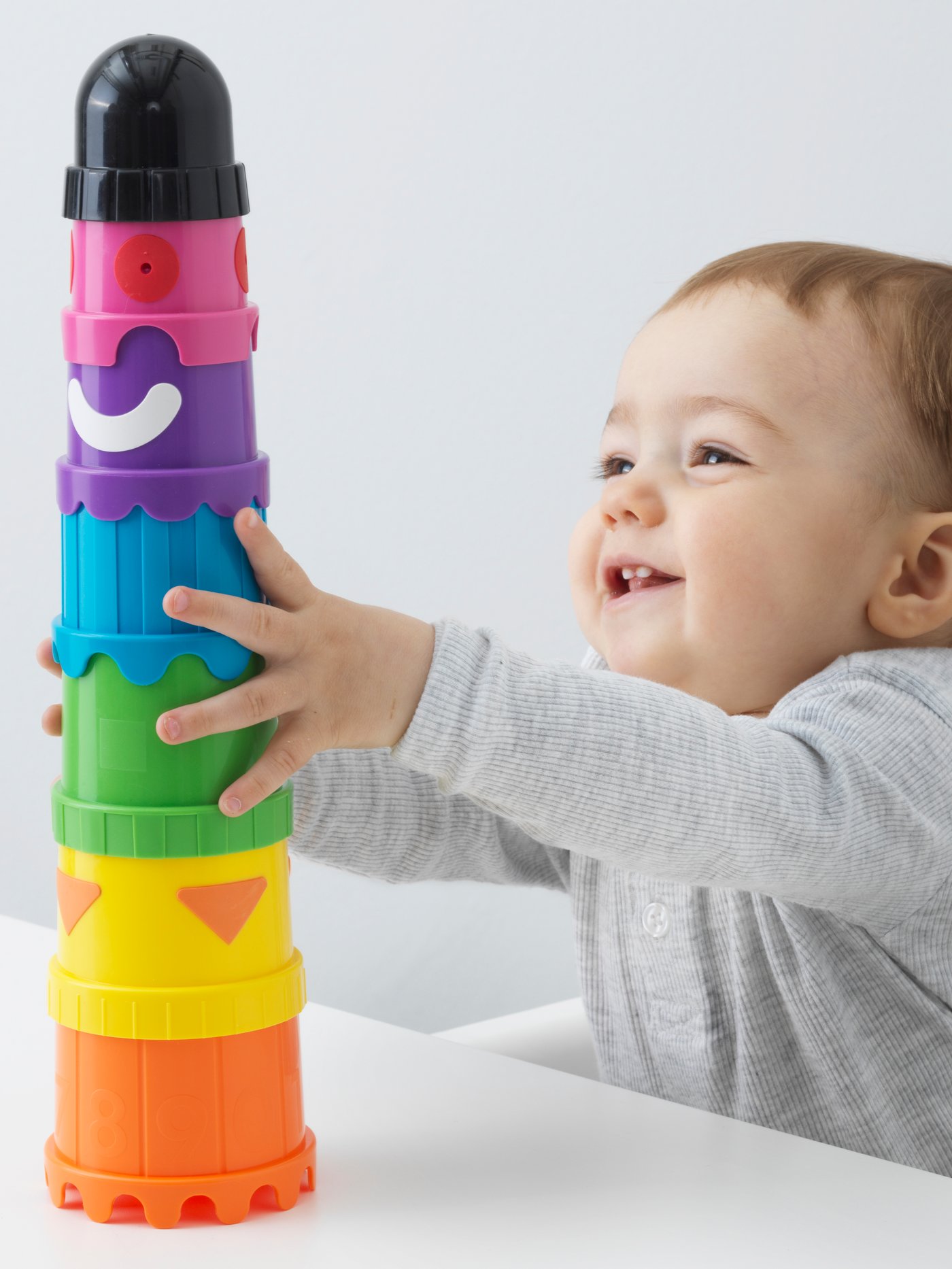 A smiling baby sits in a highchair and holds on to a tower of brightly coloured MULA building beakers on a table.