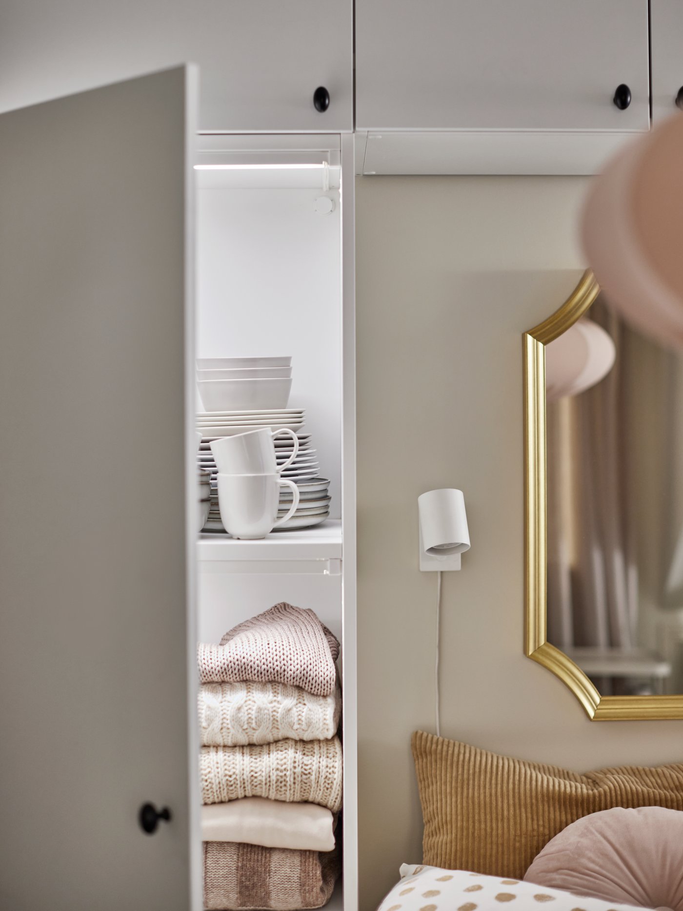 Ett vitt PLATSA garderobssystem bredvid och ovanför en säng, med en dörr öppen som visar porslin och hopvikta tröjor inuti.
