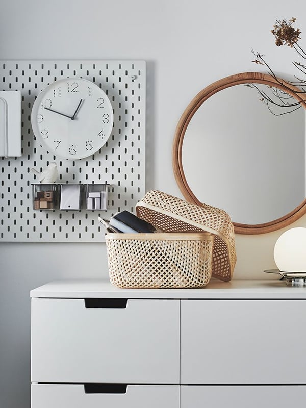 White TROMMA clock on the wall above a white NORDLI cabinet with a SMARRA rotan basket and a HINDÅS round mirror.