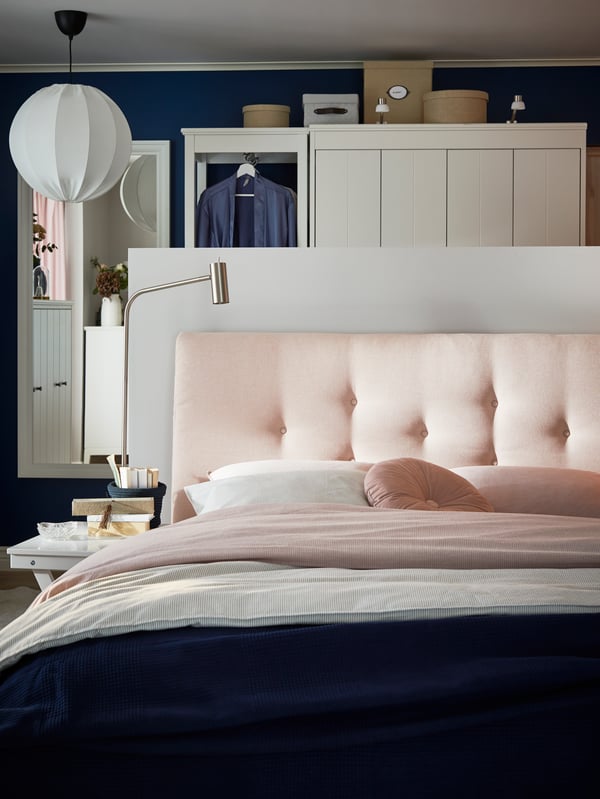 A large pink upholstered bed, pink duvet cover, blue blanket, a metal reading lamp, a white textile pendant lamp.