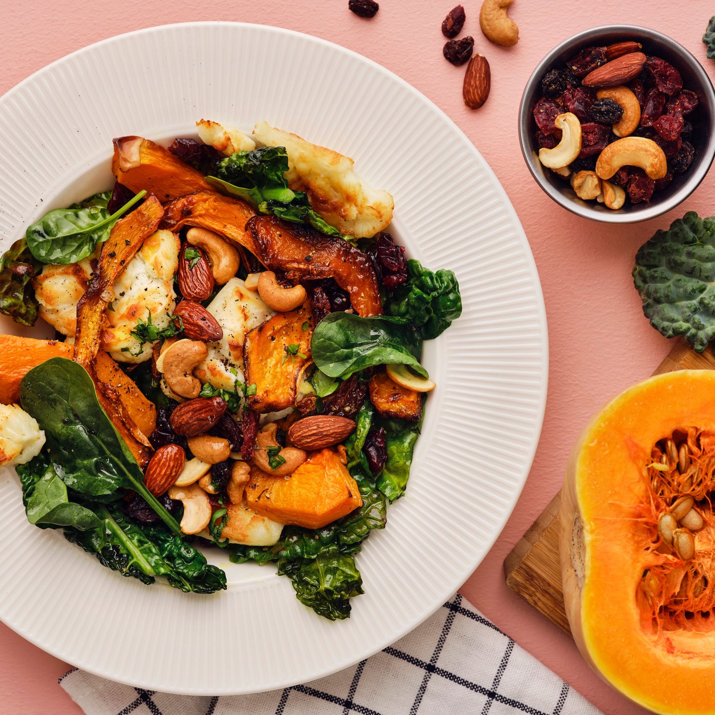 Un'insalata con zucca butternut, cavolo nero, caprino e un mix di noci tostate/bacche MUNSBIT su un tavolo rosa.