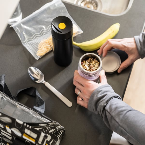 A table with diverse items, including an UNDERSÖKA insulated travel mug in black, a banana and a person holding a mug.
