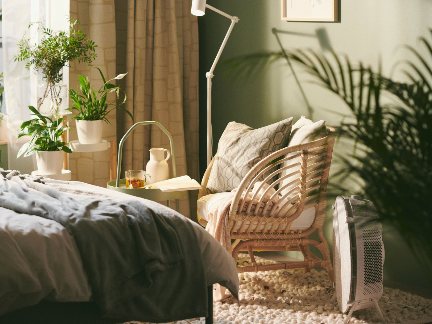 A BUSKBO rattan armchair is in a corner of a green bedroom with plenty of plants, with a STARKVIND air purifier next to it.