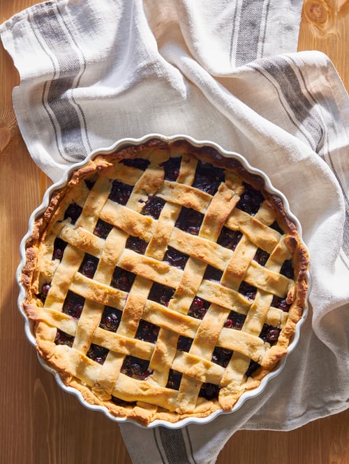Close up on kitchen counter with freshly baked fruit pie in white stoneware pie dish, on white tea towel with grey stripes.