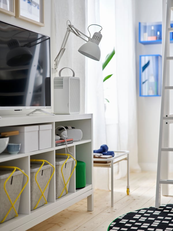 A white KALLAX TV bench with an underframe holds a VR headset, game controllers and storage baskets with a TV on top.