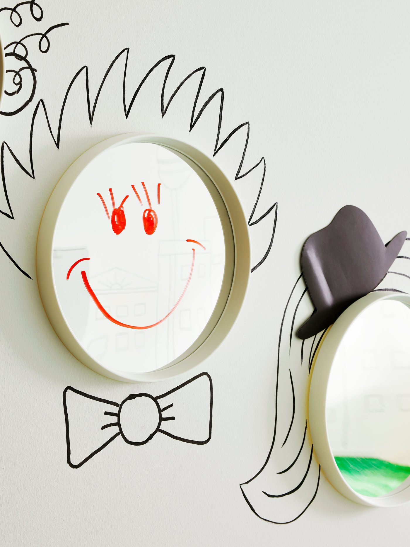 Two round HÄNGIG mirrors, one with a face drawn on it and the other with a sticker of a hat placed above it, hang on a wall.