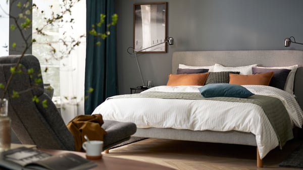 A bedroom with dark grey walls and a bed with white-grey-orange bedding, an armchair in foreground and windows in back.