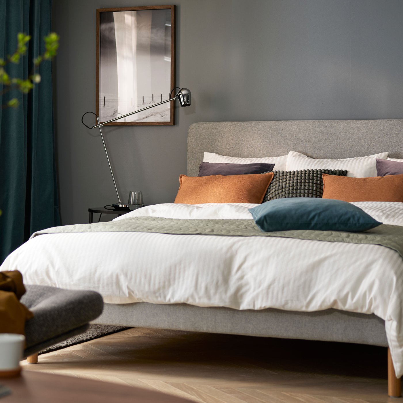 Una camera da letto con pareti grigio scuro e un letto con biancheria bianco-grigio-arancio, una poltrona in primo piano e finestre sul retro.