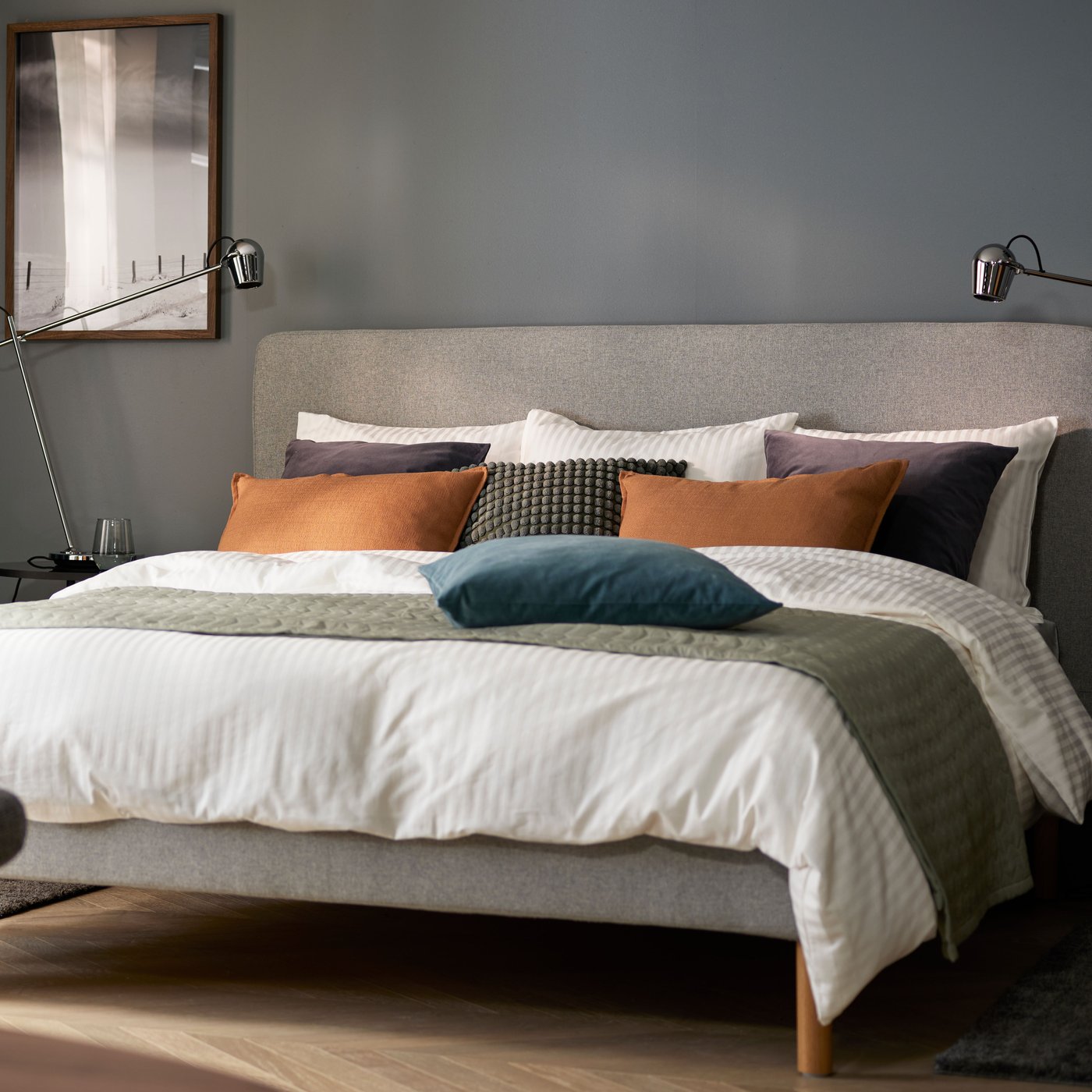 A bedroom with dark grey walls and a bed with white-grey-orange bedding, an armchair in foreground and windows in back.
