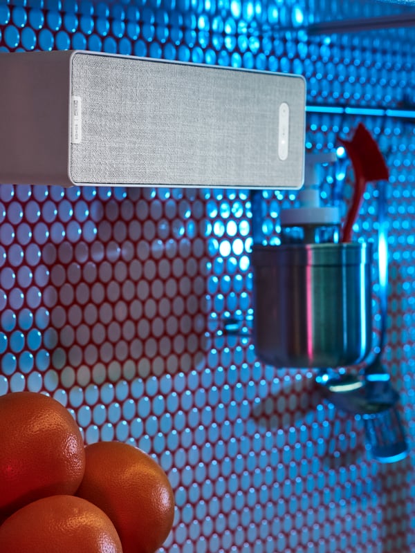 Ein weisser SYMFONISK WiFi-Regalspeaker hängt in einer Küche mit runden Fliesen an einer KUNGSFORS Stange aus Edelstahl.