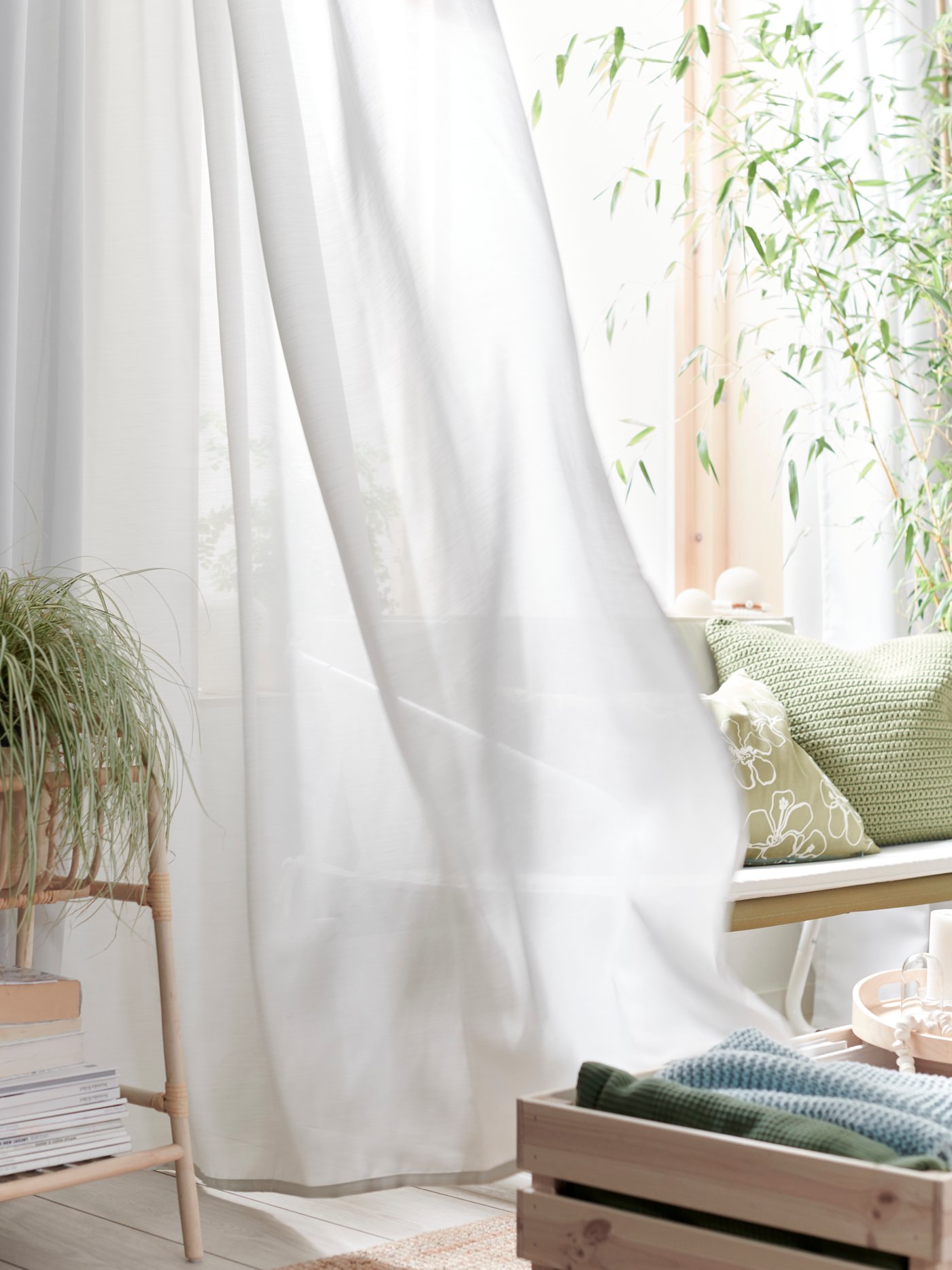 A bright living room with green plants and cushions and a light grey GUNRID air purifying curtain moving in the wind.