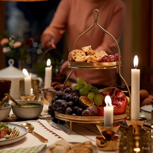 A gold-coloured GOKVÄLLÅ two-tier serving stand displays an assortment of fruits and pastries on a dining table.