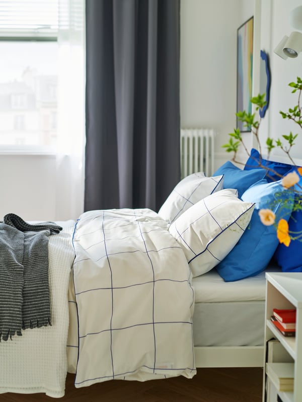 View of bed from the side with white bedding and blue pillows. Window in the background, with dark grey curtain at the side.
