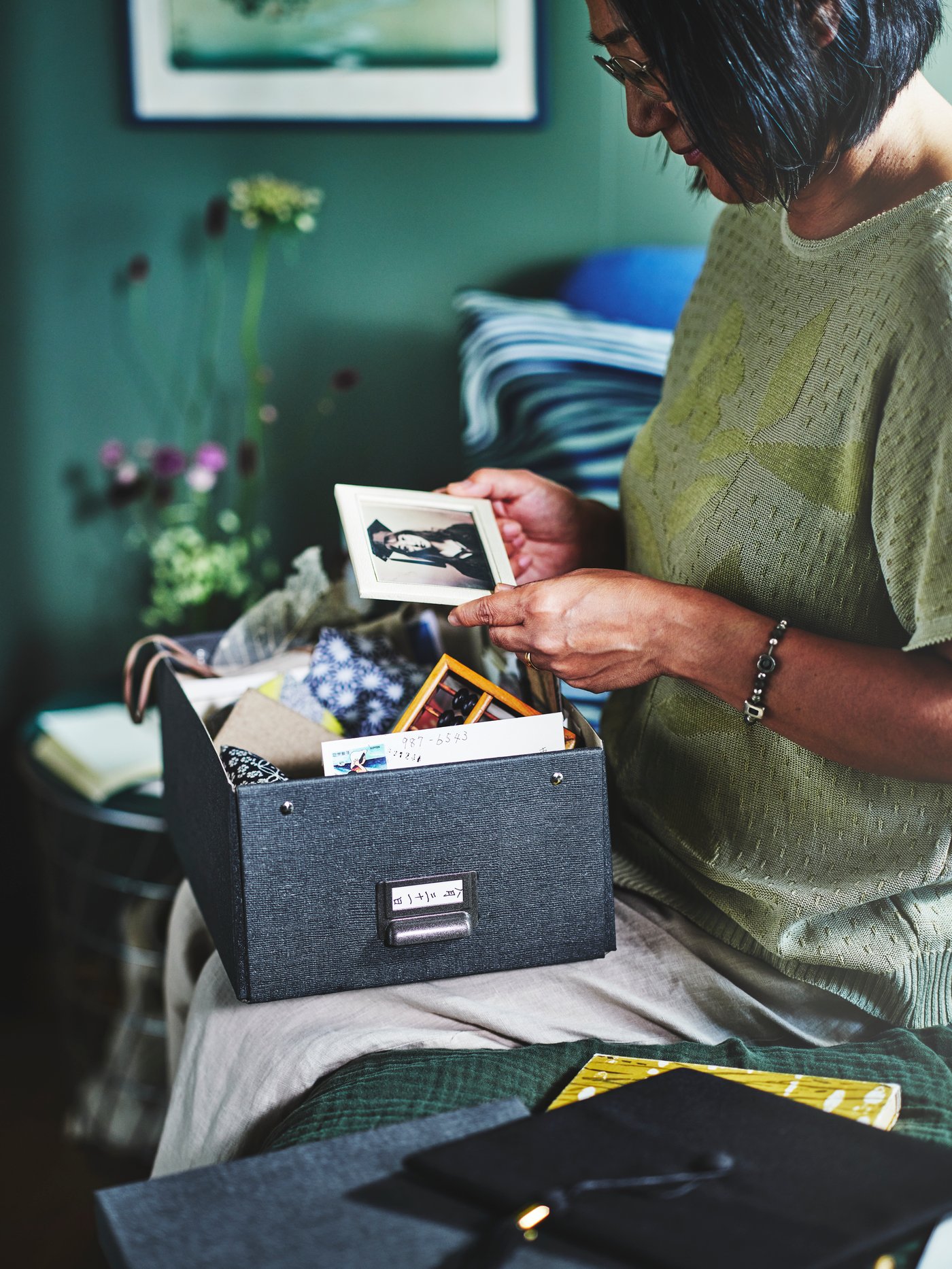 A woman on a bed with a TJOG box in her lap. She’s looking at a black and white picture of herself as a young student.