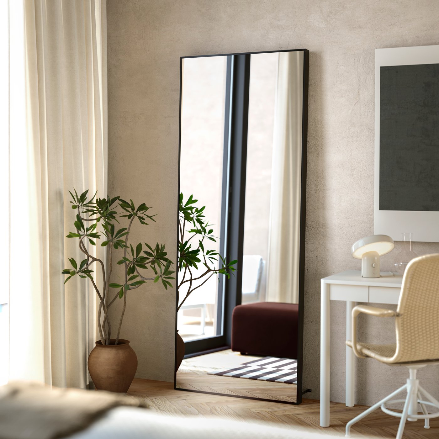 Un miroir HOVET noir pleine longueur est posé sur le sol, contre un mur blanc cassé de la chambre à coucher, reflétant la lumière naturelle.
