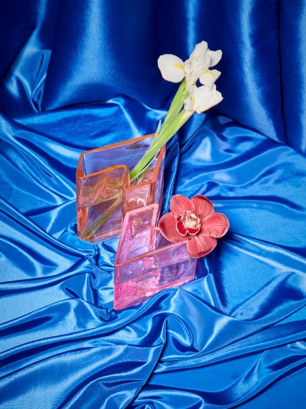 A set of two pink KARISMATISK vases are paired to make a ‘Z’ shape; the vases are against blue satin and hold flowers.