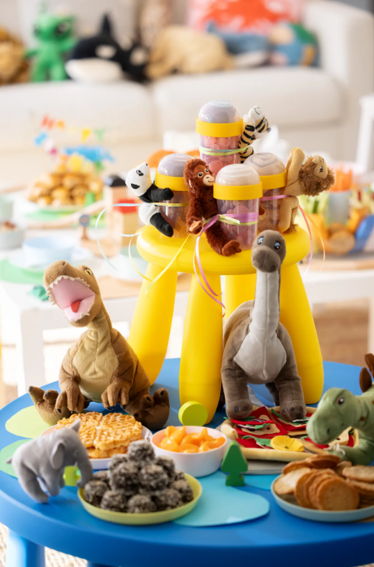 Ein MAMMUT Hocker steht als Etagere auf einem MAMMUT Tisch. Stofftierdinos bewachen die darauf angerichteten Snacks.