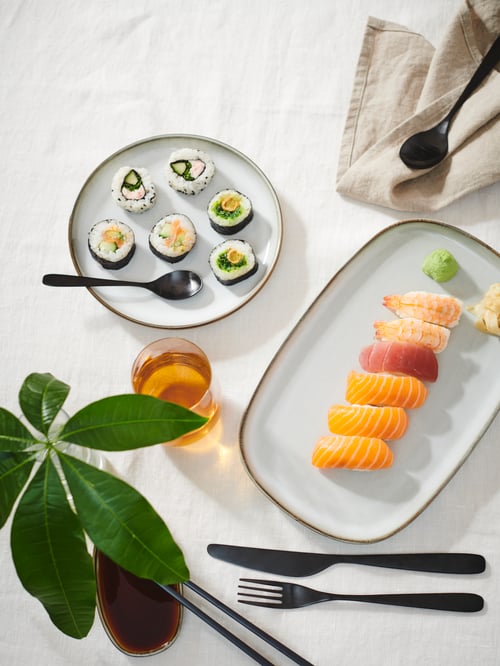Vue de dessus d’une table à manger avec un long plat de service, une assiette ronde, un ensemble de couverts noir mat, un bocal, des linges à vaisselle et une plante en pot.
