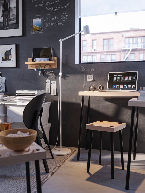A FRIDNÄS stool and two tables, and a floor lamp by a window. One table holds a laptop and headphones, the other holds books.