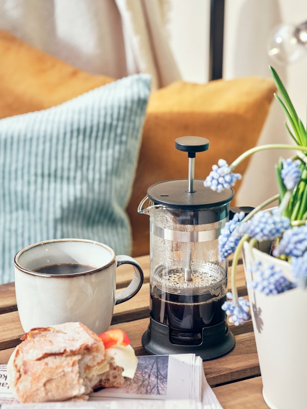 Une table d’extérieur avec une cafetière UPPHETTA, une tasse et un pot de fleurs avec des coussins bleus et jaunes en arrière-plan.