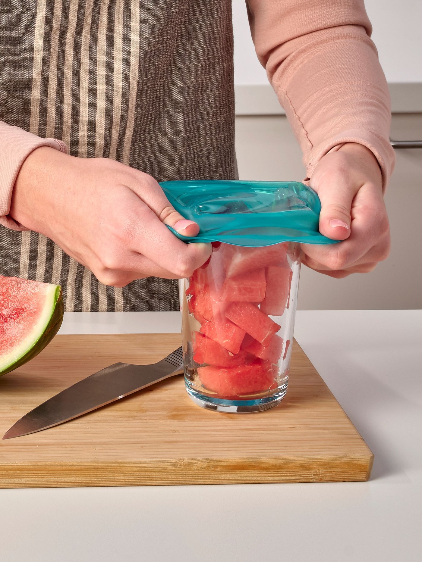 Una persona col·loca una tapa de silicona turquesa per a aliments ÖVERMÄTT sobre un got amb síndria trossejada que està sobre una taula de tallar.
