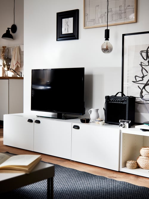 Ein Fernseher auf einer BESTÅ TV-Bank mit Türen vor einer Wohnzimmerwand mit gerahmten Bildern.