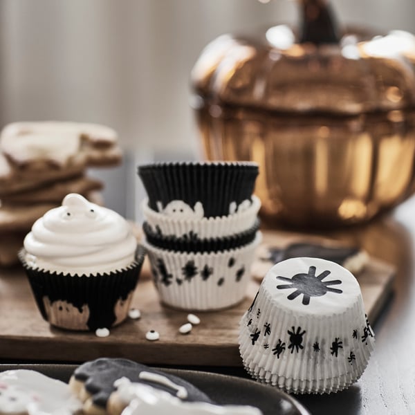 Some KUSTFYR baking cups decorated with ghosts and spiders are on a table next to a cupcake, some cookies, and a glass bowl.